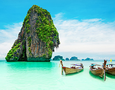 Traditional Thai boats moored on the shore of an aqua green beach with limestone cliff islands behind