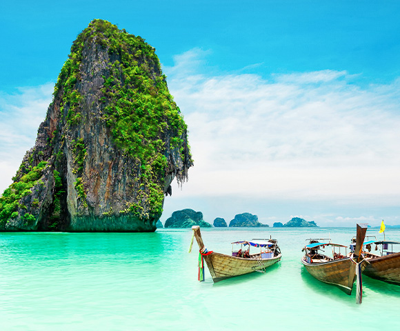 Traditional Thai boats moored on the shore of an aqua green beach with limestone cliff islands behind