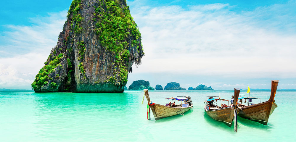 Traditional Thai boats moored on the shore of an aqua green beach with limestone cliff islands behind