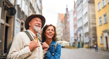 A smiling elderly couple stands on a cobblestone street lined with colorful historic buildings. They appear joyful and relaxed, enjoying a sunny day.