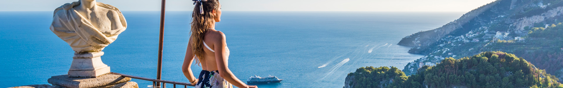 A woman in a floral dress stands on a balcony, overlooking the serene sea and mountains. A marble bust statue is beside her under a bright blue sky.