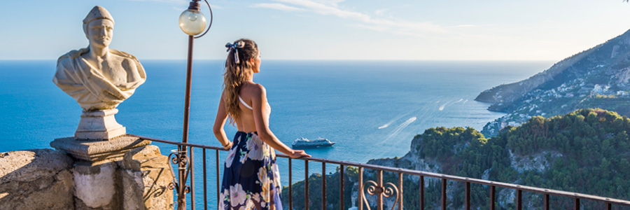 A woman in a floral dress stands on a balcony, overlooking the serene sea and mountains. A marble bust statue is beside her under a bright blue sky.