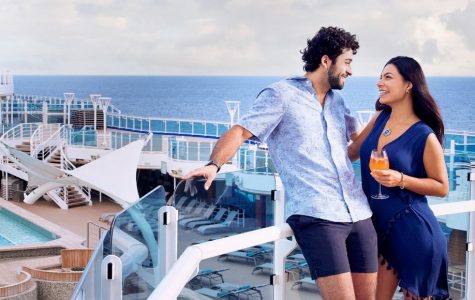 Smiling couple enjoying drinks on a sunny cruise ship deck, with lounge chairs, glass railings, and the blue ocean behind them.