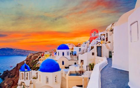 Panoramic Santorini coastline at sunset, with whitewashed cliffside buildings, blue domes, a vivid blue window, and calm Aegean Sea.