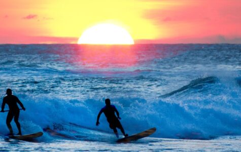 Two surfers riding waves at sunset with a large sun near the horizon.
