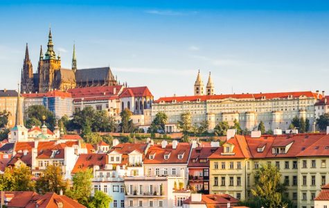 Sunny panoramic view of Prague across the Vltava River, with red-roofed historic buildings, Prague Castle, and St. Vitus Cathedral.