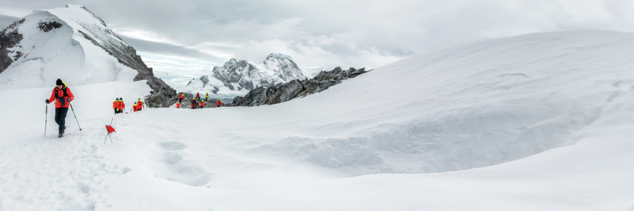 A group of hikers in bright orange jackets climbs a snow-covered mountain with trekking poles. Rugged peaks are visible in the background under a cloudy sky.