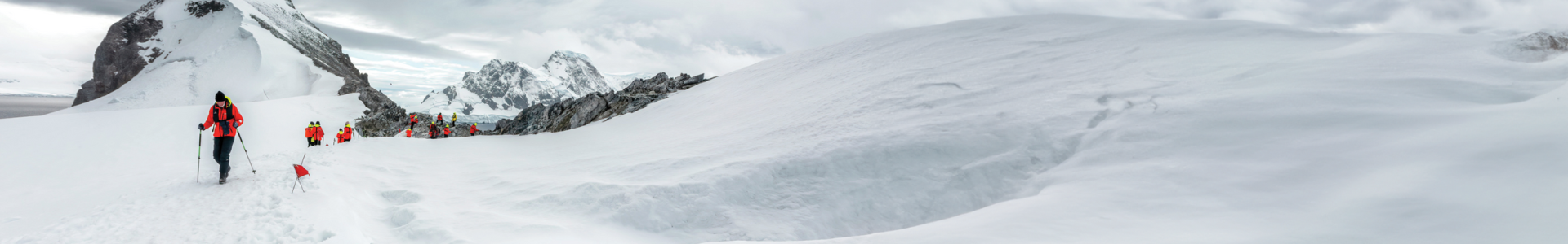 A group of hikers in bright orange jackets climbs a snow-covered mountain with trekking poles. Rugged peaks are visible in the background under a cloudy sky.