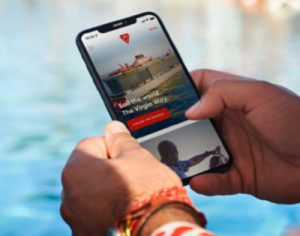 A person holds a smartphone displaying a cruise line website by a pool. The screen shows an image of a ship with the text "Sail the world, the Virgin way."