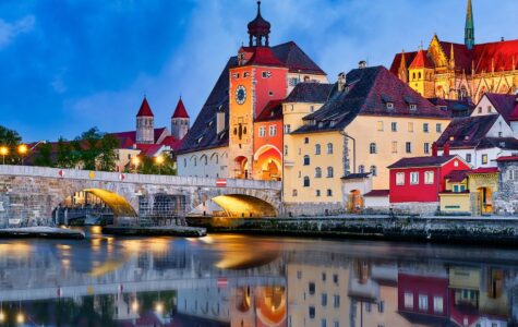 Evening view of an illuminated Gothic cathedral and stone bridge with reflections on the water.