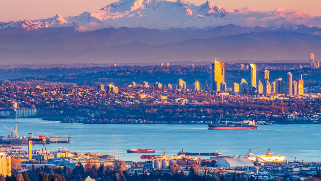 City skyline at sunrise with a mountain backdrop. Sunlit buildings and ships on a calm blue river create a serene, picturesque scene.