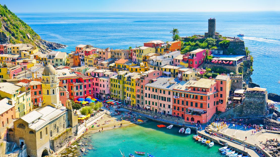 Colorful seaside village with pastel buildings overlooking a turquoise harbor, dotted with small boats. A sunny, lively Mediterranean atmosphere.