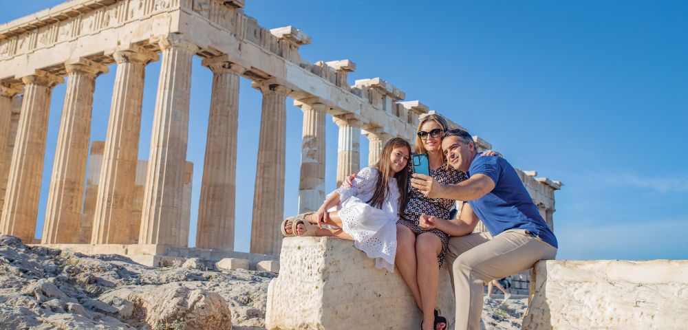 A family takes a selfie in front of the Parthenon in Athens. The mother, father, and daughter smile warmly under a clear blue sky, evoking joy and adventure.