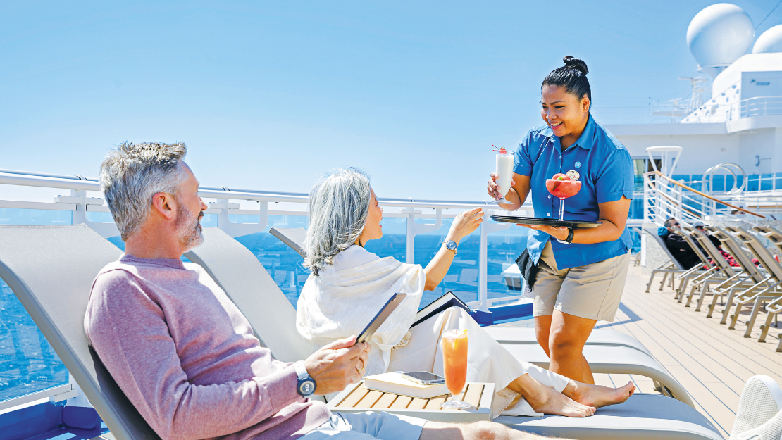 A cruise ship deck scene with lounge chairs, a server delivering colorful cocktails, bright sunshine, and a scenic ocean backdrop.