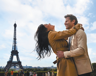 A joyful couple embraces in front of the Eiffel Tower, surrounded by onlookers under a partly cloudy sky.