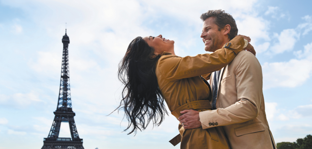 A joyful couple embraces in front of the Eiffel Tower, surrounded by onlookers under a partly cloudy sky.