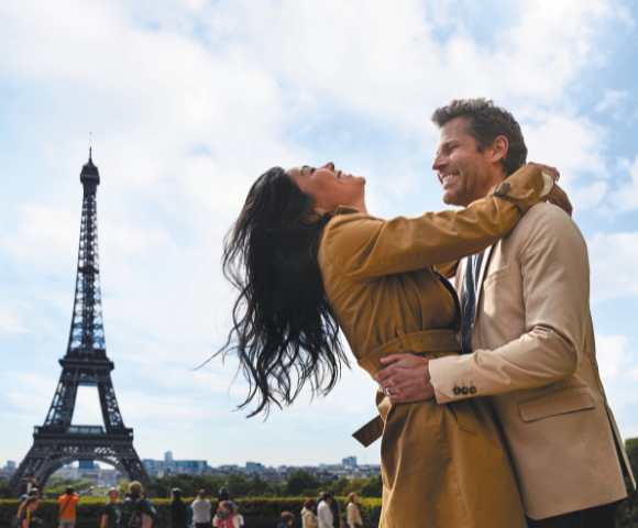 A joyful couple embraces in front of the Eiffel Tower, surrounded by onlookers under a partly cloudy sky.