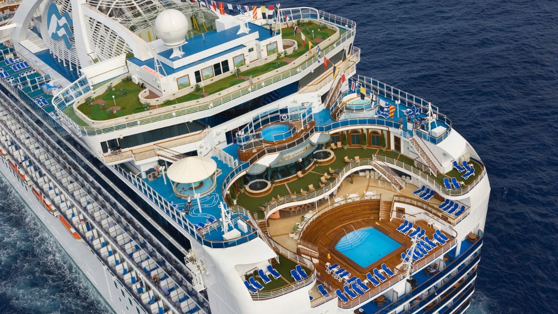 Aerial view of a cruise ship deck featuring pools, lounge chairs, and gardens, set against a deep blue ocean background.