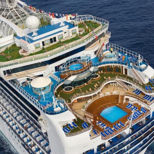Aerial view of a cruise ship deck featuring pools, lounge chairs, and gardens, set against a deep blue ocean background.