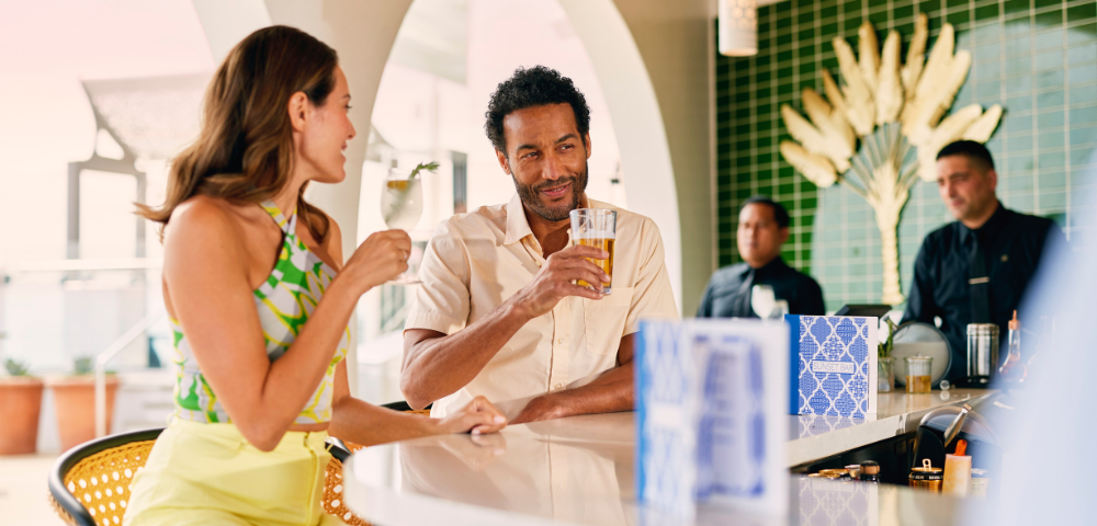 A stylish bar scene features two people enjoying drinks, surrounded by attentive staff and vibrant decor in a sunny, tropical atmosphere.