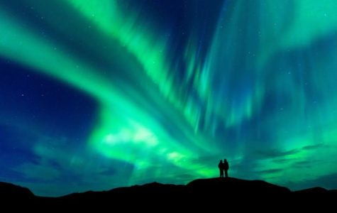 A couple stands on a hill, silhouetted against the vibrant aurora borealis lighting up the night sky.