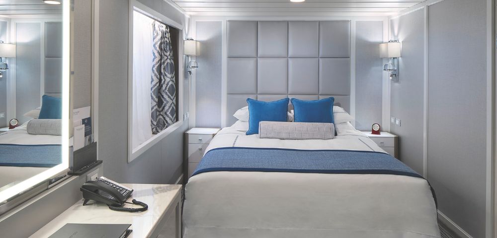 Modern stateroom featuring a plush bed with blue accents, gray walls, and a mirror with soft lighting onboard the Oceania Regatta.