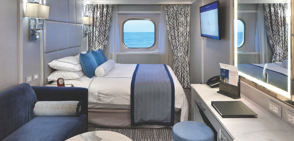 Modern cruise stateroom featuring a plush bed, ocean view window, modern décor, and a cozy seating area. Bright and inviting atmosphere onboard Oceania Regatta.