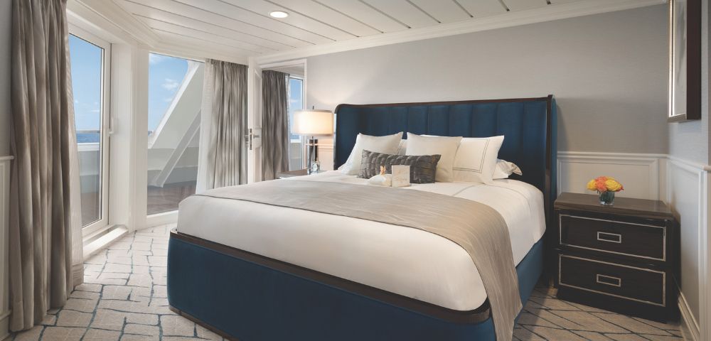 A modern cruise stateroom featuring a large plush bed, luxurious linens, large balcony windows, and stylish decor with a lamp and nightstand onboard Oceania Regatta.