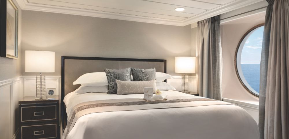 Cosy stateroom onboard Regatta featuring a plush bed, bedside lamps, and a circular window with ocean views. Elegant decor in light tones.
