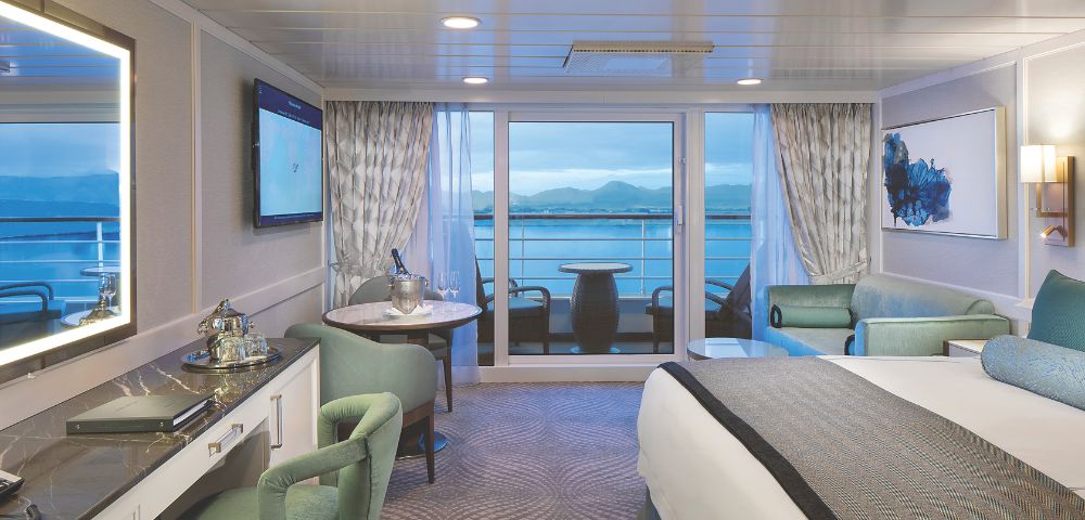 Luxurious cruise ship cabin onboard Regatta featuring a plush bed, modern decor, a seating area and a balcony with ocean views.