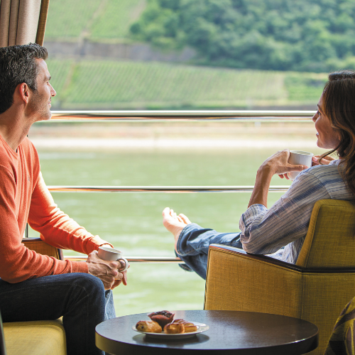 A woman sits relaxed in a chair, sipping from a cup, while a man looks out at the serene river view through large windows.