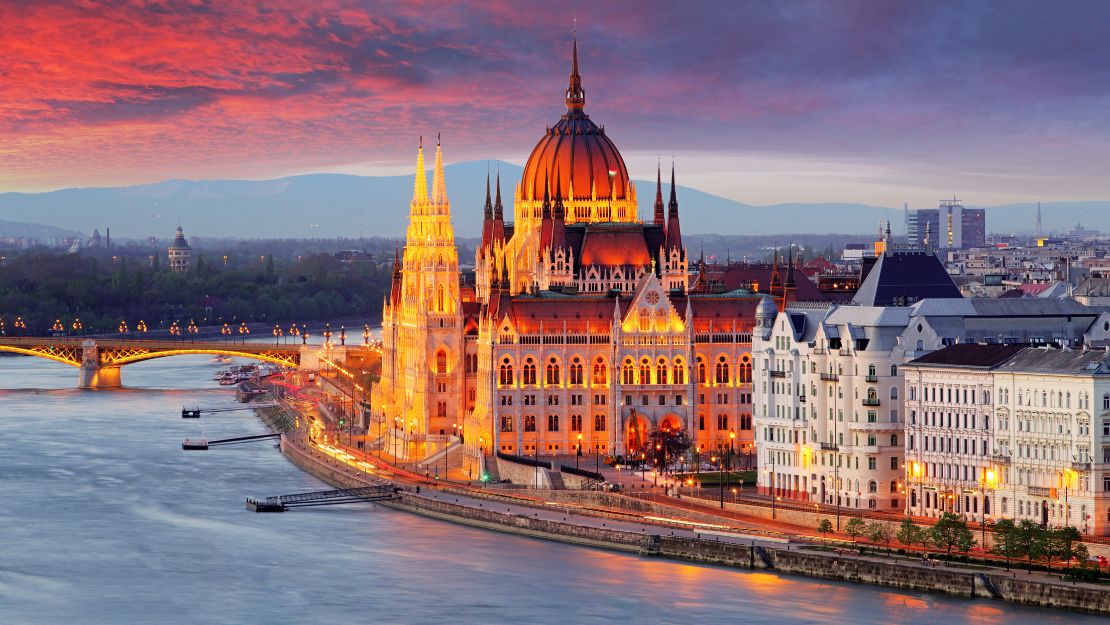 The illuminated Hungarian Parliament Building at dusk, alongside the Danube River, with dramatic clouds in the sky.