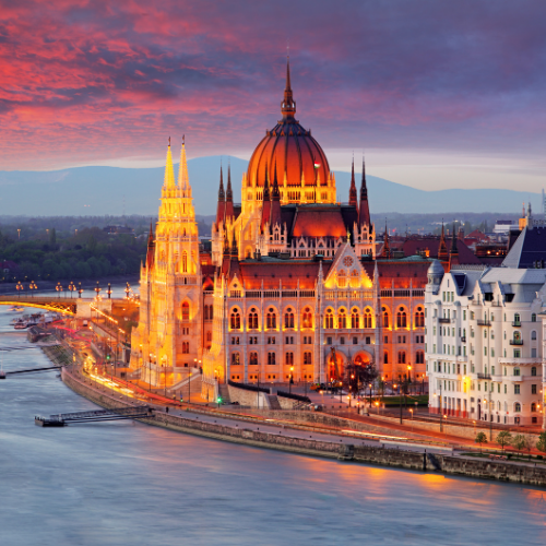 The illuminated Hungarian Parliament Building at dusk, alongside the Danube River, with dramatic clouds in the sky.