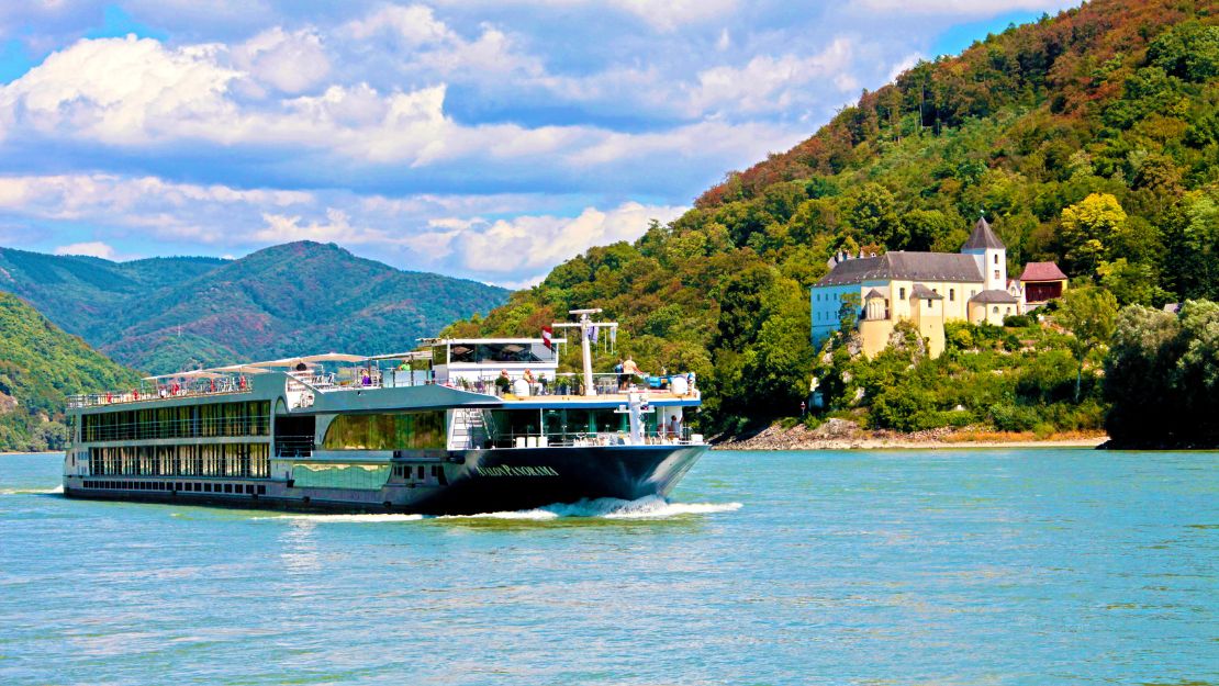 A river cruise ship glides through calm waters, with lush green hills and a historic castle-like building on the riverbank.
