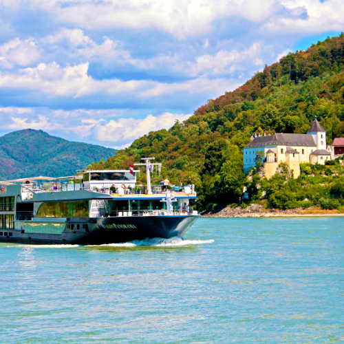 A river cruise ship glides through calm waters, with lush green hills and a historic castle-like building on the riverbank.