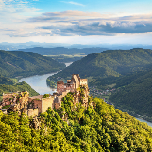 A scenic view of a medieval castle perched on a rocky cliff, surrounded by lush green forests and a winding river below, under a blue sky.