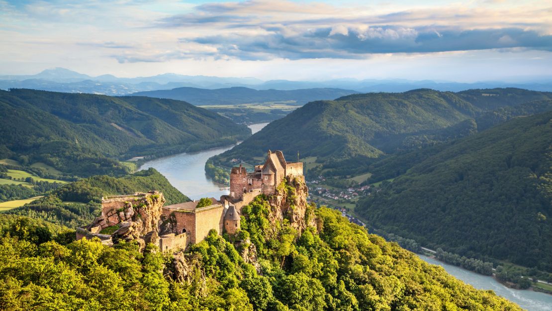A river cruise ship glides through calm waters, with lush green hills and a historic castle-like building on the riverbank.