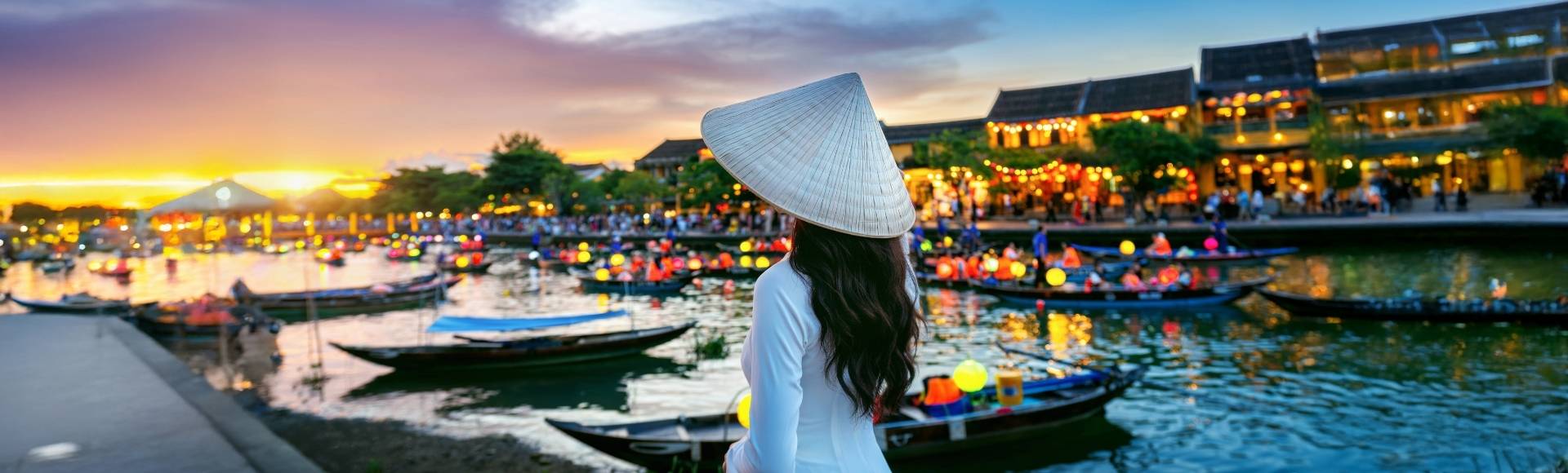 A woman stands by a river at sunset, with colorful lanterns and boats reflecting in the water.