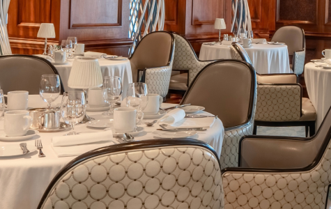 An elegant dining room with cream-clothed tables, patterned chairs, tall curtained windows and a glowing oval ceiling onboard Regatta.
