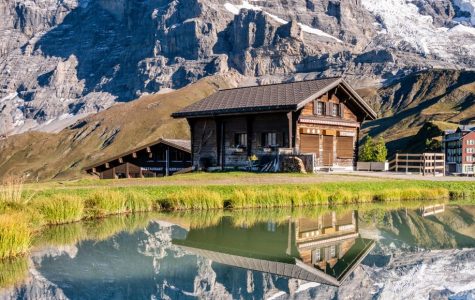 Wooden cabins sit by a tranquil pond, reflecting snow-capped mountains under a clear blue sky. The scene evokes serenity and natural beauty.