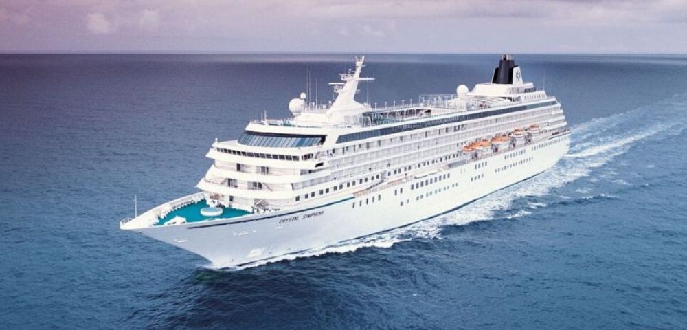 A large white cruise ship sails through calm blue waters, creating gentle waves. The ship's decks are visible, conveying a sense of luxury and leisure