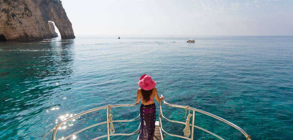 A woman in a pink hat stands at the edge of a boat, gazing at a calm blue sea and distant cliffs under a partly cloudy sky, evoking serenity and wanderlust.