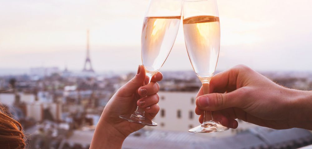 Two hands clink champagne glasses against a hazy Paris skyline at sunset, with the Eiffel Tower subtly visible in the background, evoking romance and celebration.