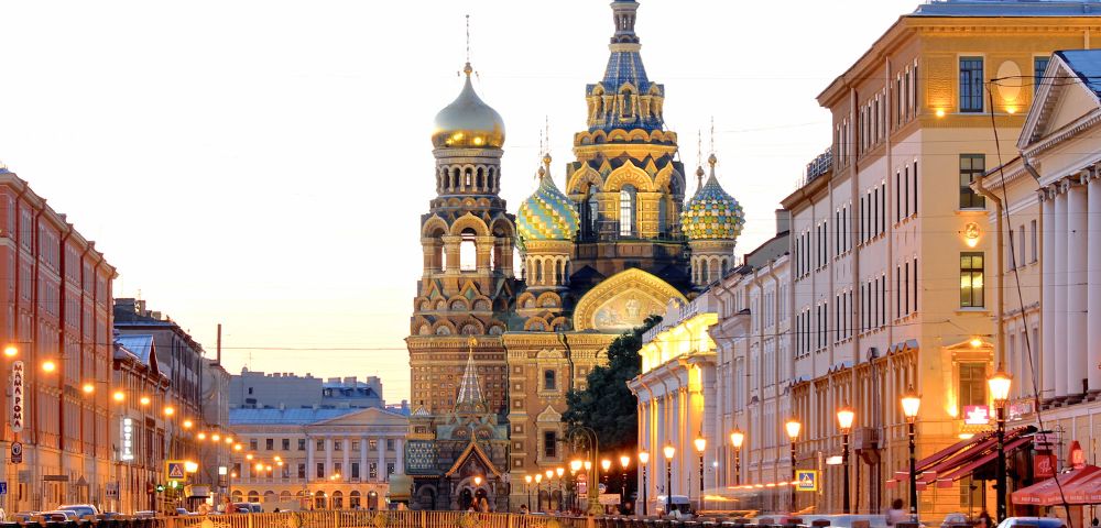 Evening view of the Church of the Savior on Spilled Blood in St. Petersburg, Russia. Lit street lamps reflect in the canal, creating a warm, serene atmosphere.