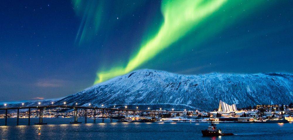 Cityscape under a night sky with vibrant green Northern Lights. Snowy mountain, illuminated bridge, and a boat on the water create a serene winter scene.