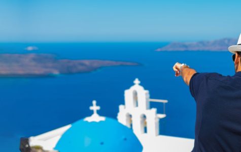 A couple wearing hats sits on a wall overlooking the sea, with white buildings and a blue-domed church in Santorini, Greece, in the background.