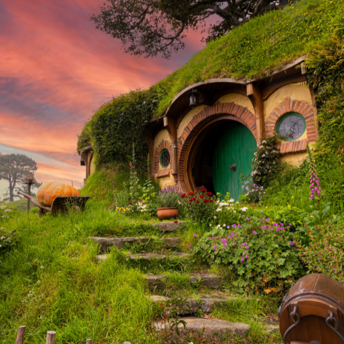 Cozy hobbit house built into a grassy hillside with a round green door, surrounded by colorful flowers under a pink sunset sky.