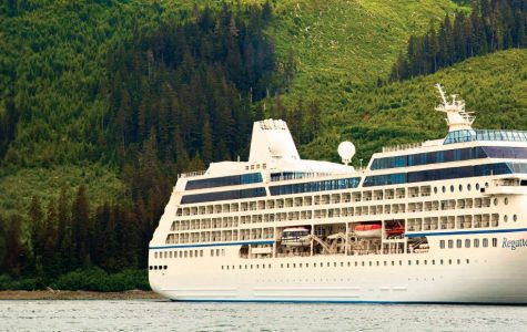 A large white cruise ship named "Regatta" sails in calm water against a backdrop of dense, lush green forest covering steep hills.