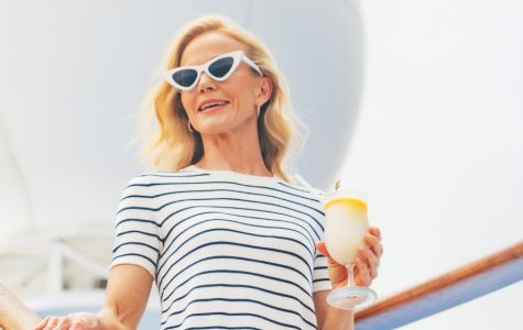 Smiling blonde woman in sunglasses and a striped shirt holds a drink on a bright ship deck, framed by curved railings under a sunny sky.