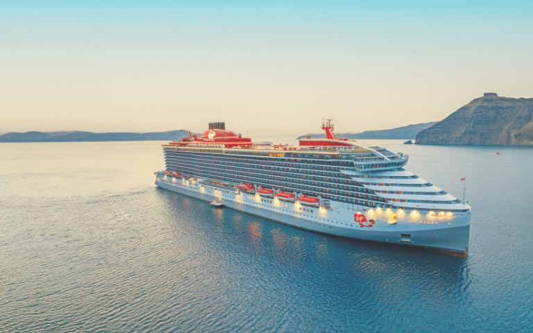 A large white Virgin Voyages cruise ship with red accents sails on calm blue water near rocky cliffs during a soft, hazy sunrise or sunset light.
